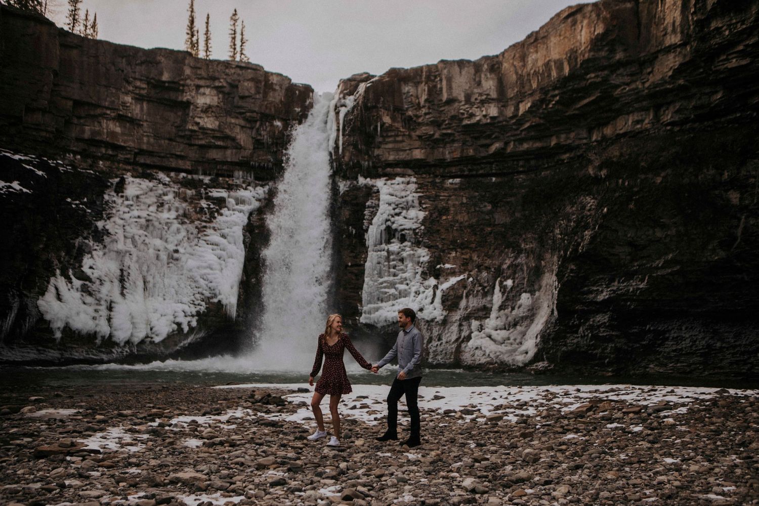 A couple dances together on rocky terrain beneath a towering waterfall.
