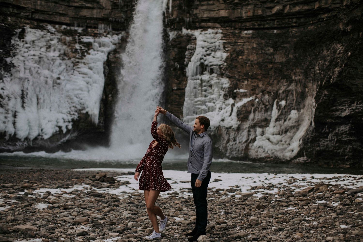 A couple dances playfully in front of a dramatic waterfall cascading down rocky cliffs.