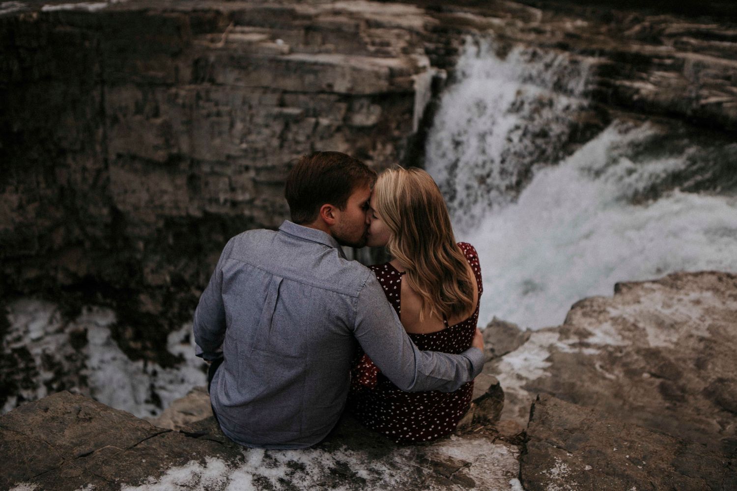 Couple sitting close together on rocky overlook with waterfall in background.