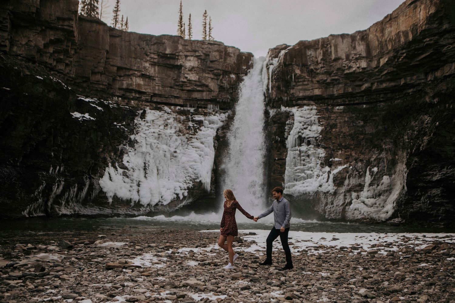 Two silhouettes hold hands while standing before a powerful waterfall in a rocky canyon.
