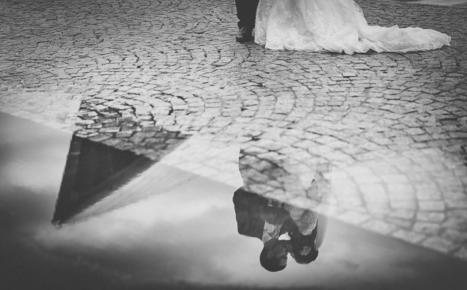 Black and white reflection of bride and groom on cobblestone street captured in a puddle.