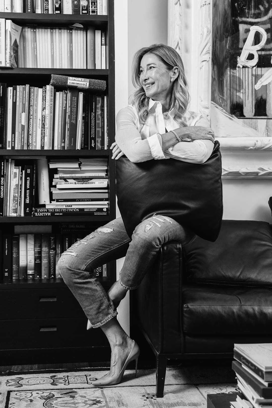 Black and white photo of a lady sitting casually on a couch by a bookshelf, barefoot and holding a large pillow.