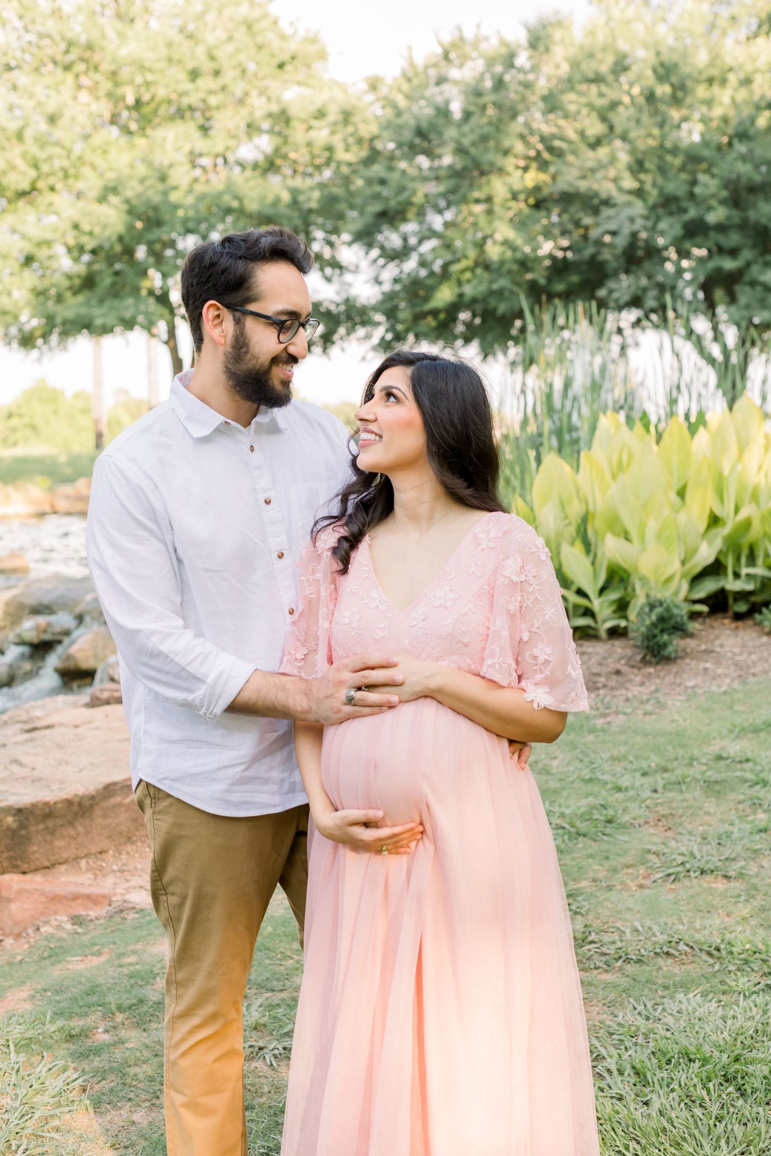 A couple smiles at each other while standing in a scenic outdoor setting with greenery.