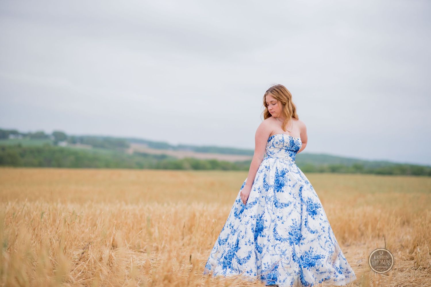 Person wearing a strapless blue and white patterned gown stands elegantly in a harvested field landscape.