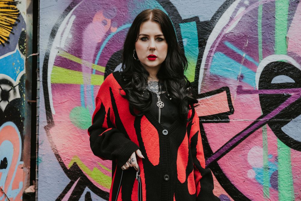 Flo an alternative model  wearing a red and black cardigan looking at camera with a graffiti wall behind shot in Hove
