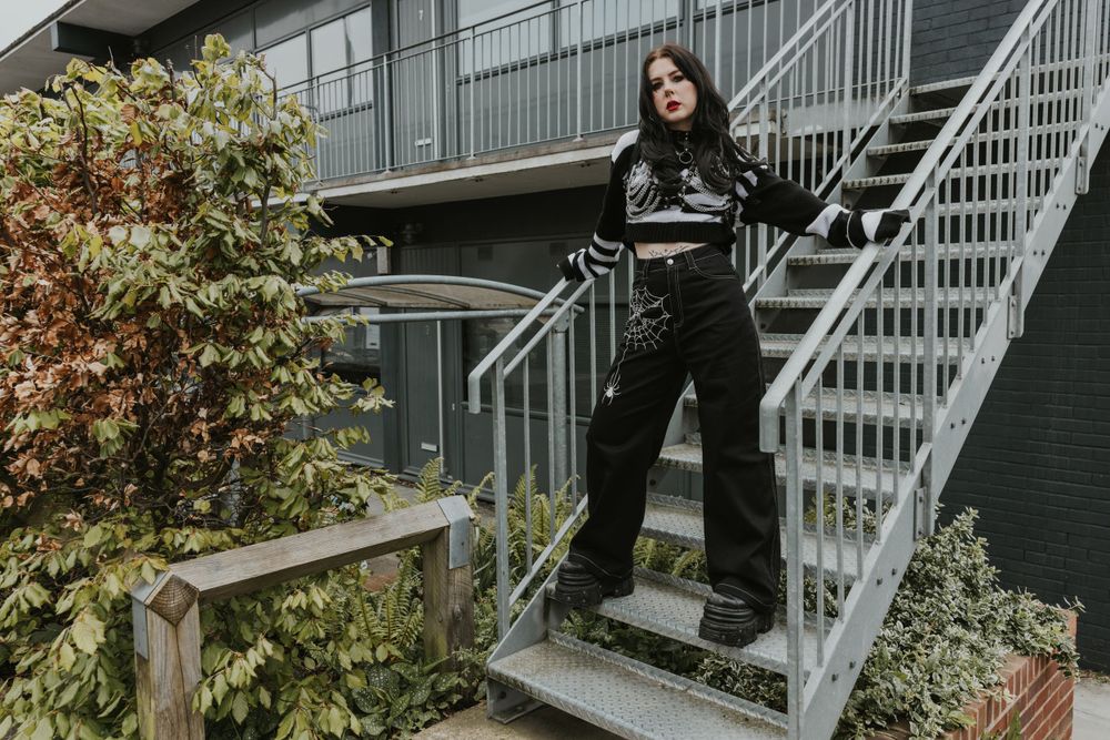 Flo an alternative model with black long hair and tattoos wearing black alternative clothing on an industrial site