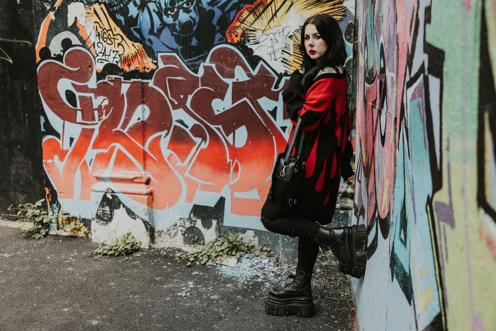 Flo an alternative model  wearing a red and black cardigan looking at camera with a graffiti wall behind shot in Hove
