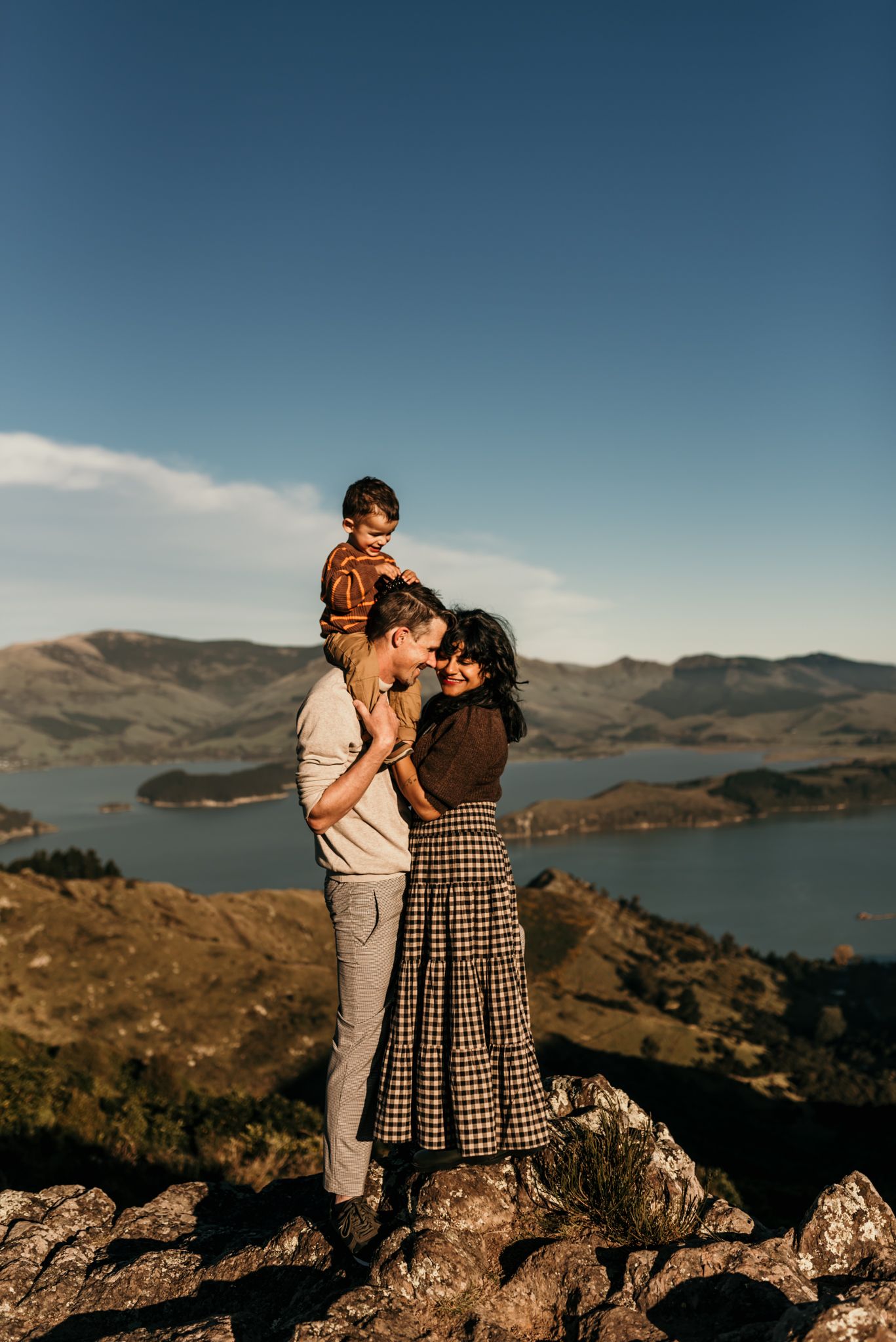 Family shares intimate moment while standing on rocky overlook with scenic mountain lake backdrop.