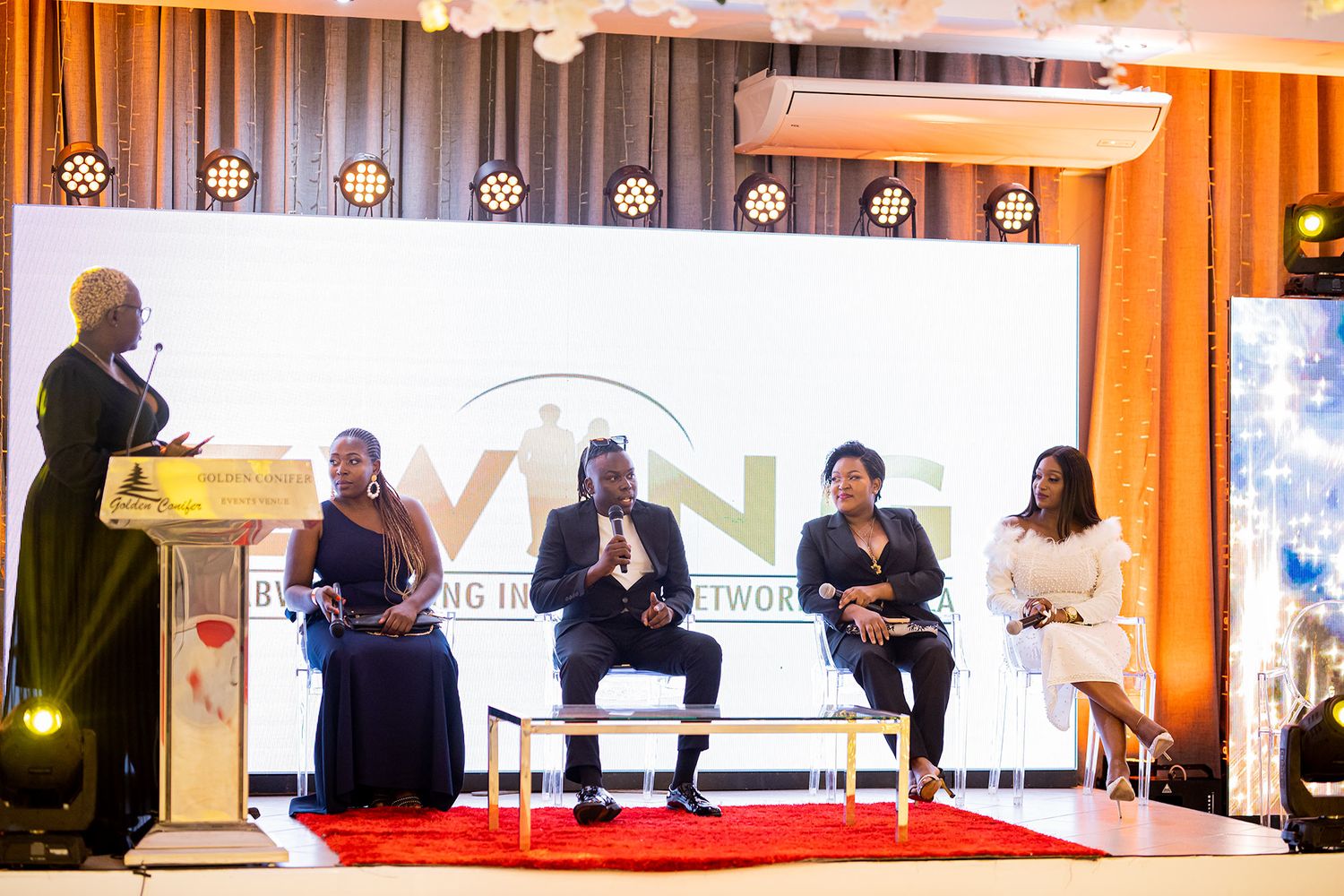 Business panel discussion setup with speakers seated on stage with orange backdrop and lighting.