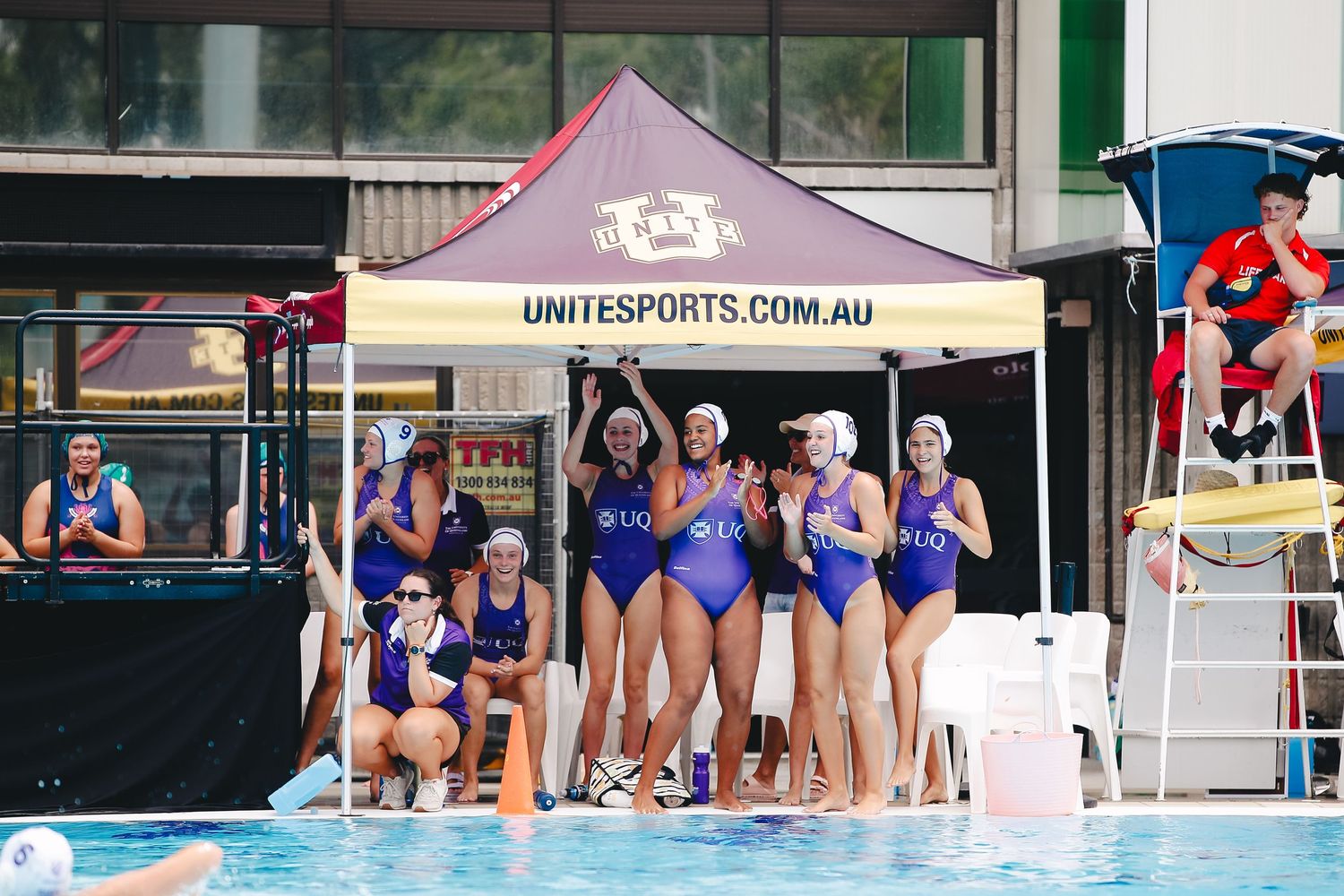 Team in blue swimsuits huddles together poolside under a sports tent during competition.