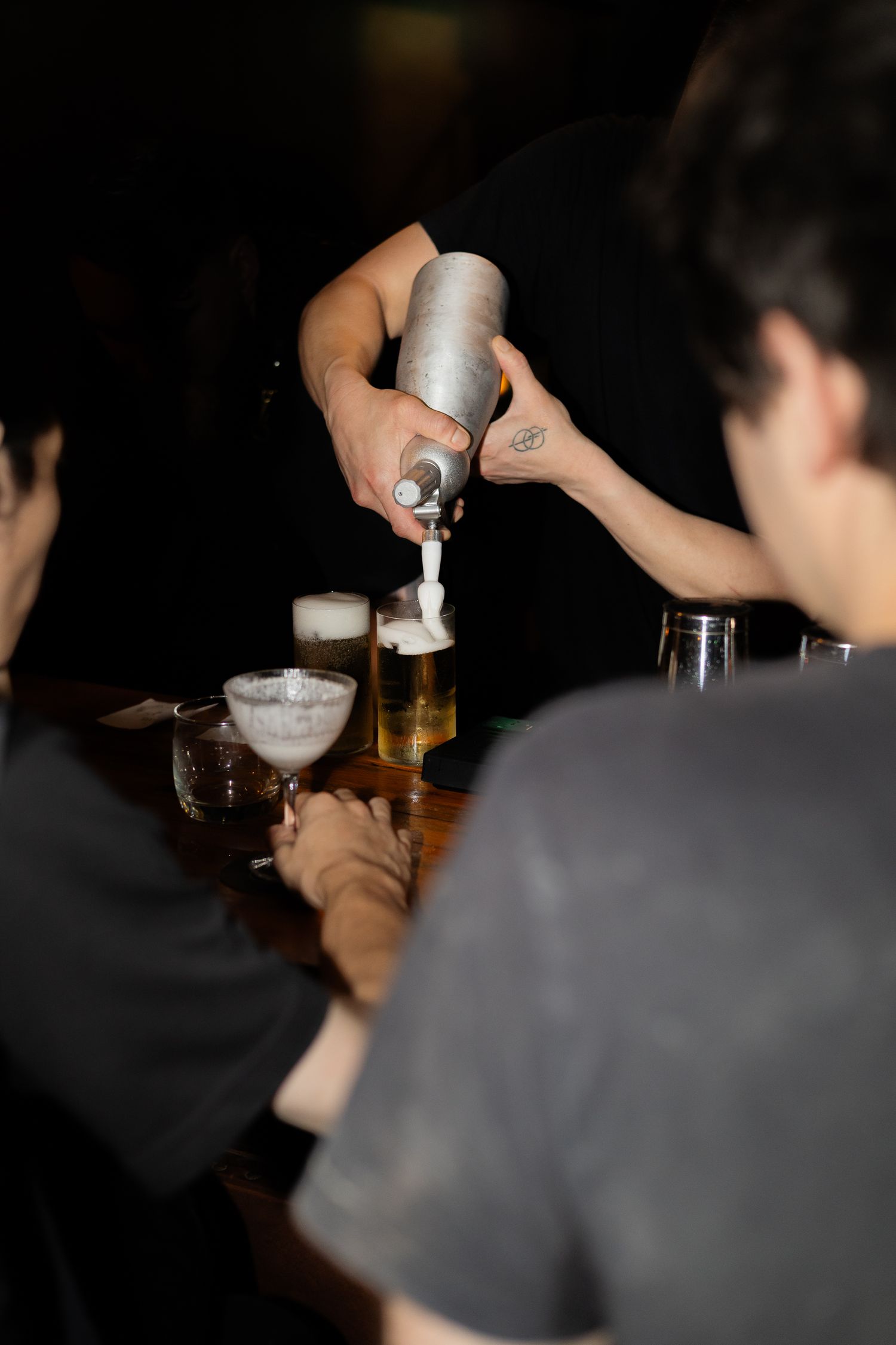 Hand pouring a cocktail with a silver shaker into martini glasses at a dimly lit bar.