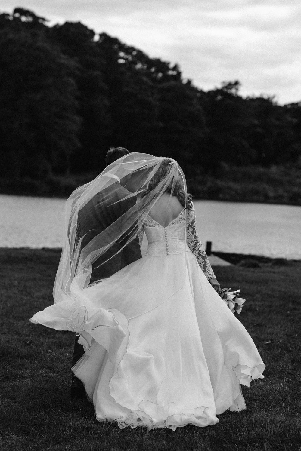 White wedding dress flowing in the wind on grassy field near lake.