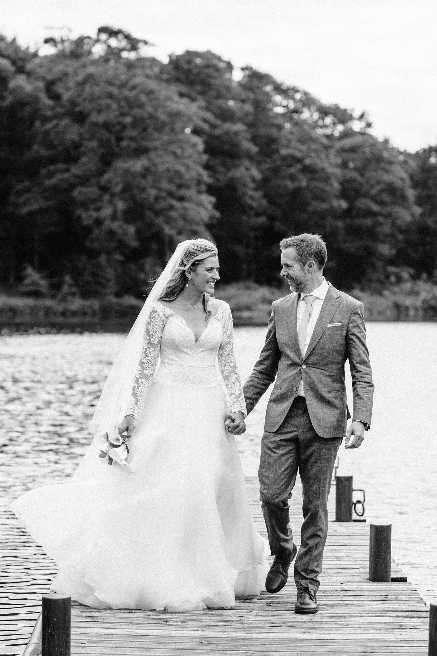 Wedding couple walks hand in hand along lakeside dock with flowing white dress and grey suit.