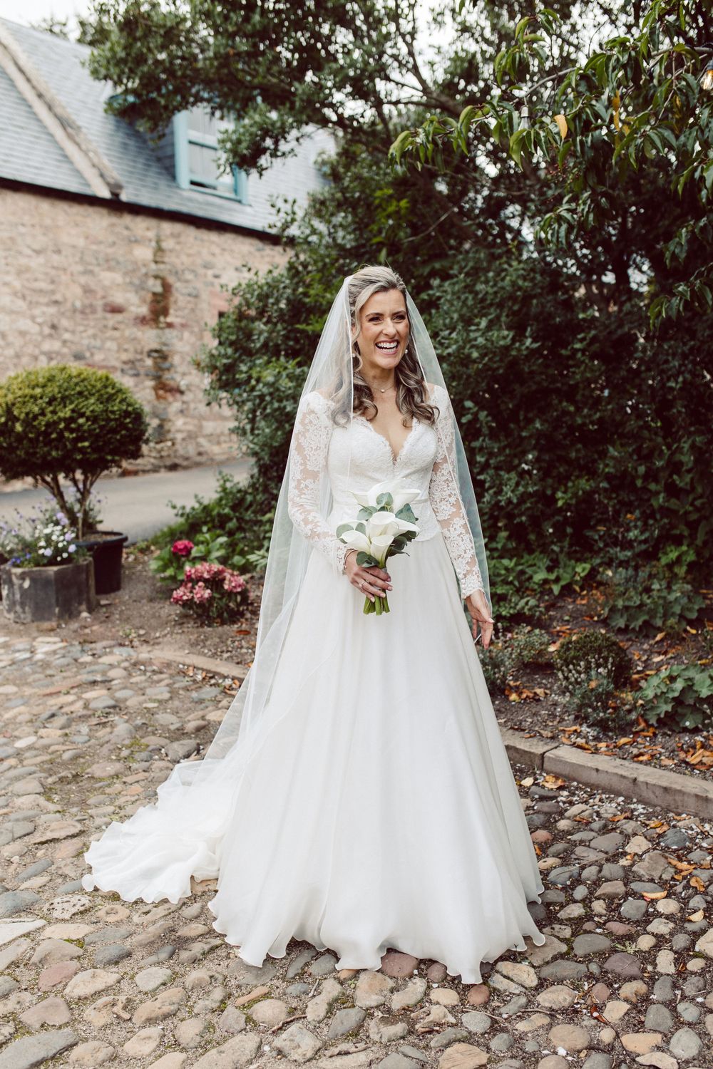 Bride in elegant white gown poses on cobblestone path surrounded by greenery.