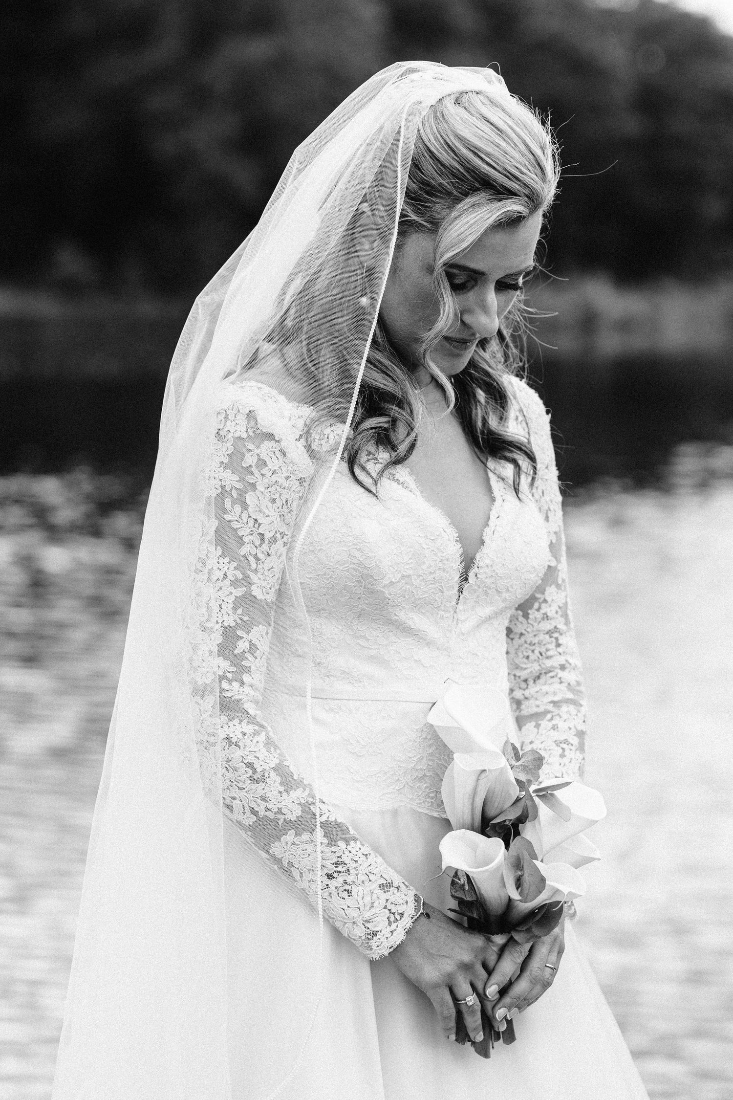 Bride in lace sleeve wedding dress and long veil poses by lake.
