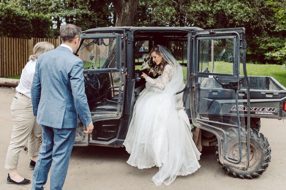Wedding party gathers around vintage car during outdoor celebration.