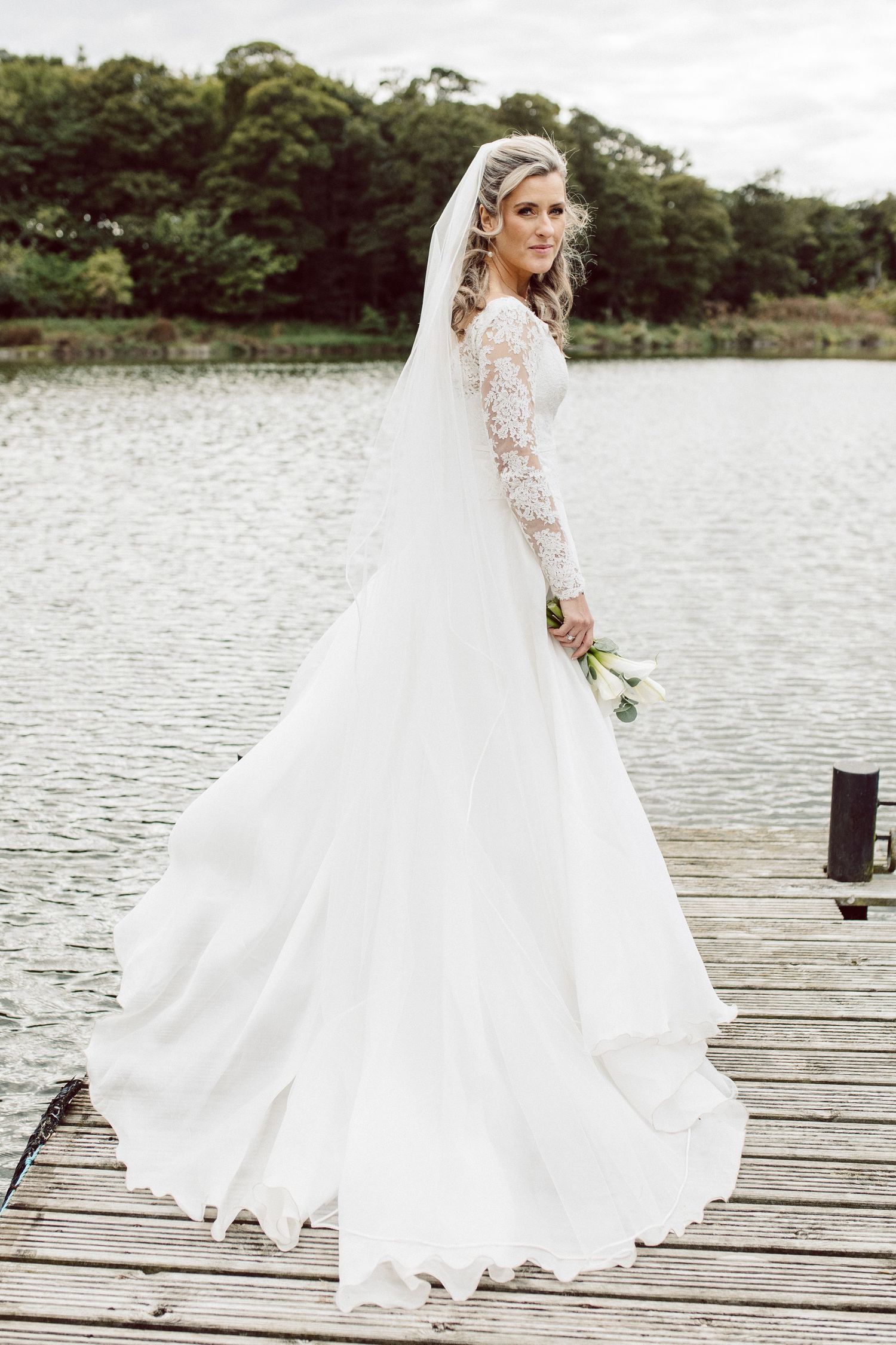 Elegant white wedding dress with lace sleeves flows beautifully on wooden dock by serene lake.