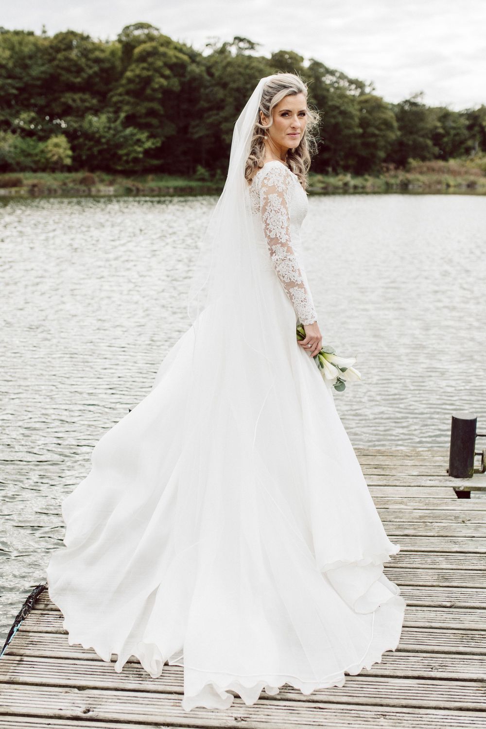 Elegant white wedding dress with lace sleeves flows beautifully on wooden dock by serene lake.
