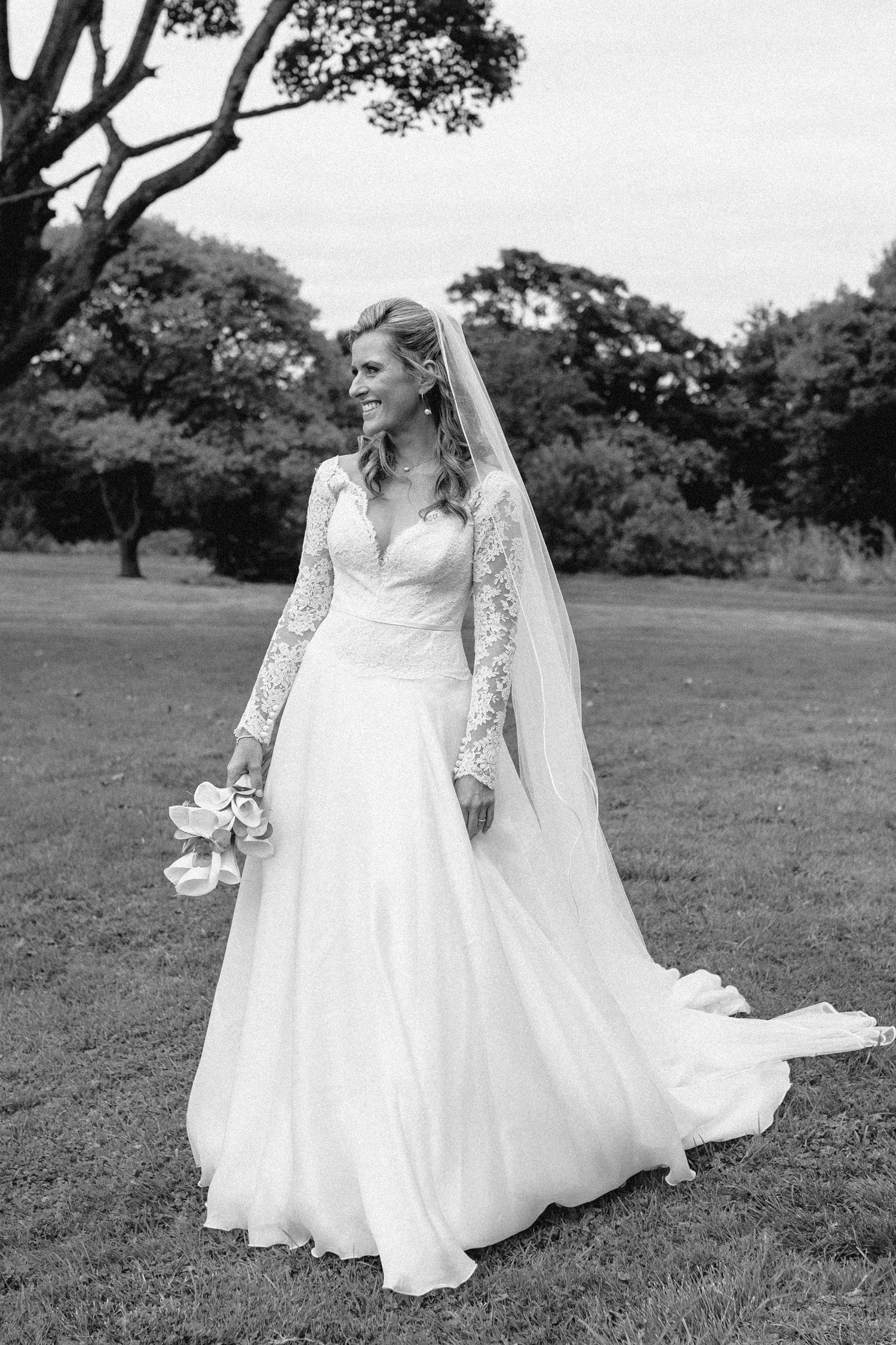 Classic bridal portrait in flowing white gown with lace sleeves against pastoral landscape.