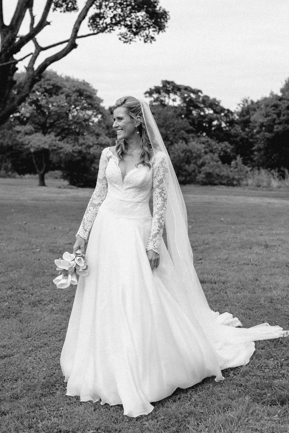 Classic bridal portrait in flowing white gown with lace sleeves against pastoral landscape.