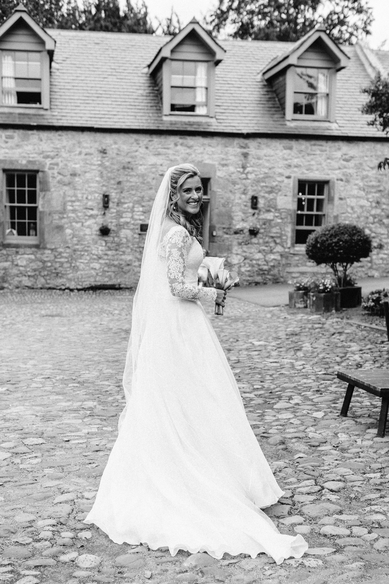Black and white photo series of bride in flowing white dress walking on cobblestone path by stone building.