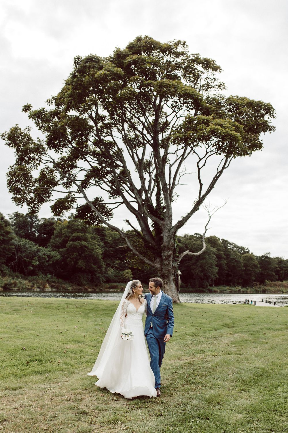 Romantic wedding couple strolls through green meadow with majestic trees in background.
