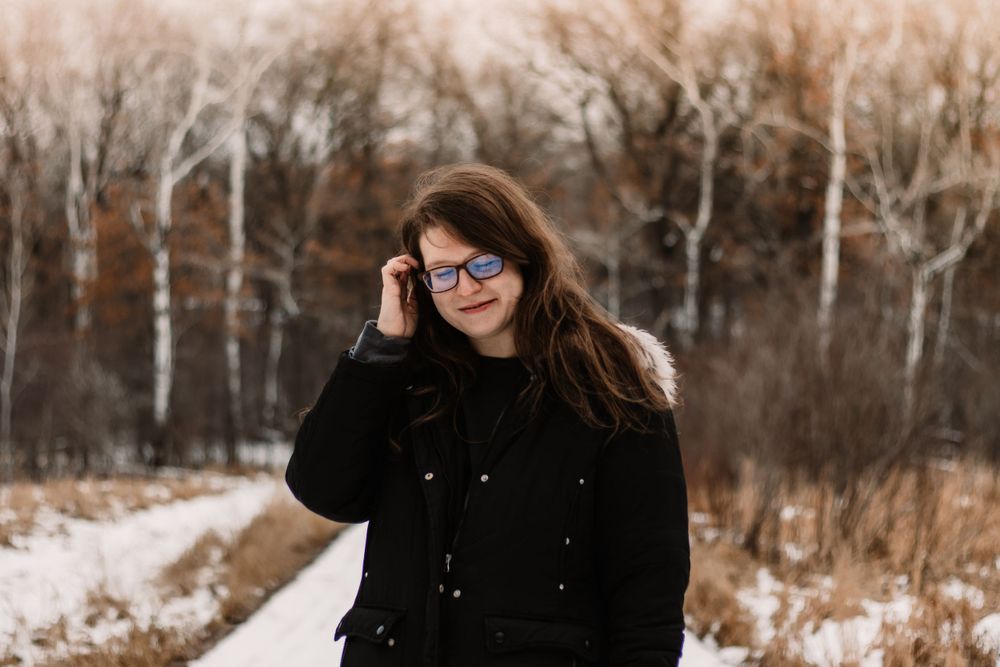 Raimi Ellen tucking her hair behind her ear in front of a snowy forest trail background