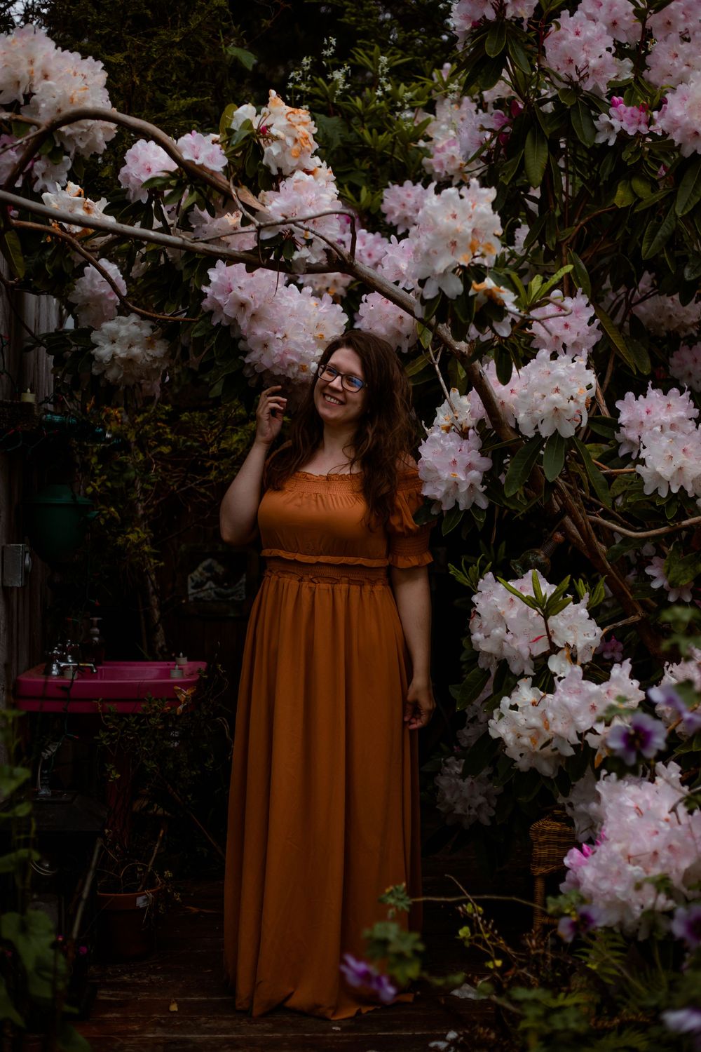 Raimi Ellen in an orange dress with pink flowers and green leaves surrounding her as she stands smiling at the camera