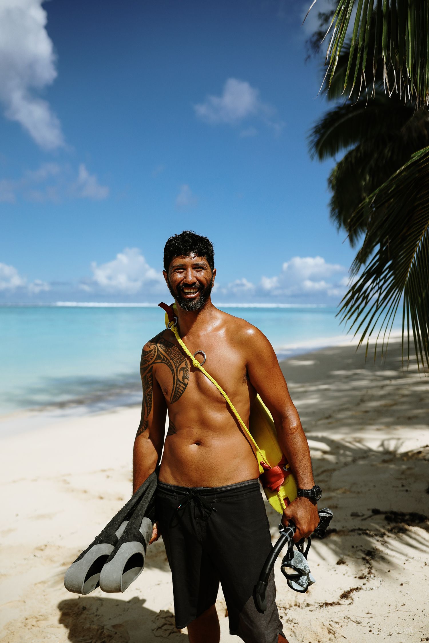 Beachgoer with tattoo and snorkel gear smiles on white sand beach under palm trees.