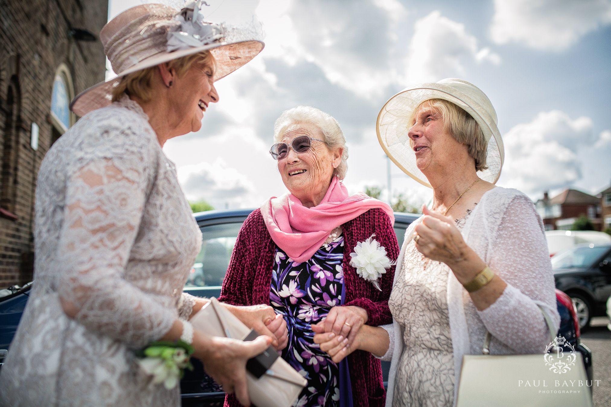 Three elegant ladies in formal wear and hats sharing a laugh at outdoor event.
