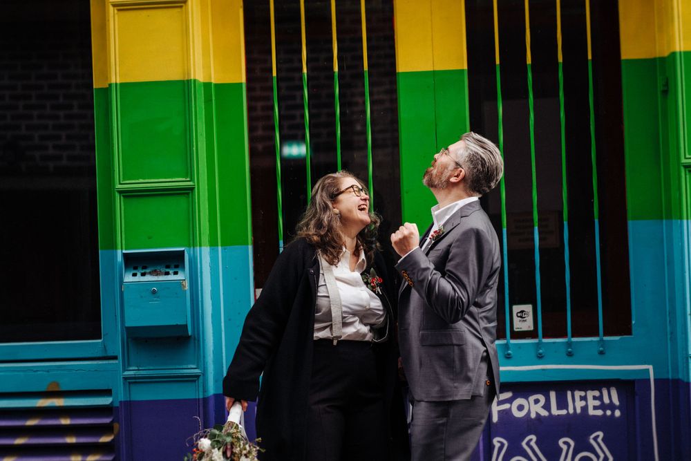 A couple shares a playful moment in front of a vibrant rainbow-colored building.