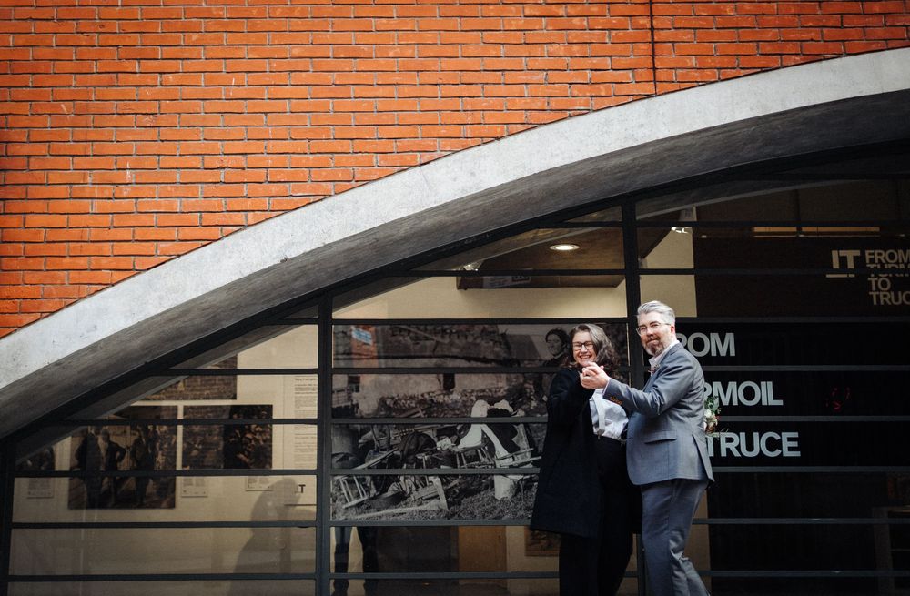 A couple poses beneath a curved architectural feature against red brick walls.