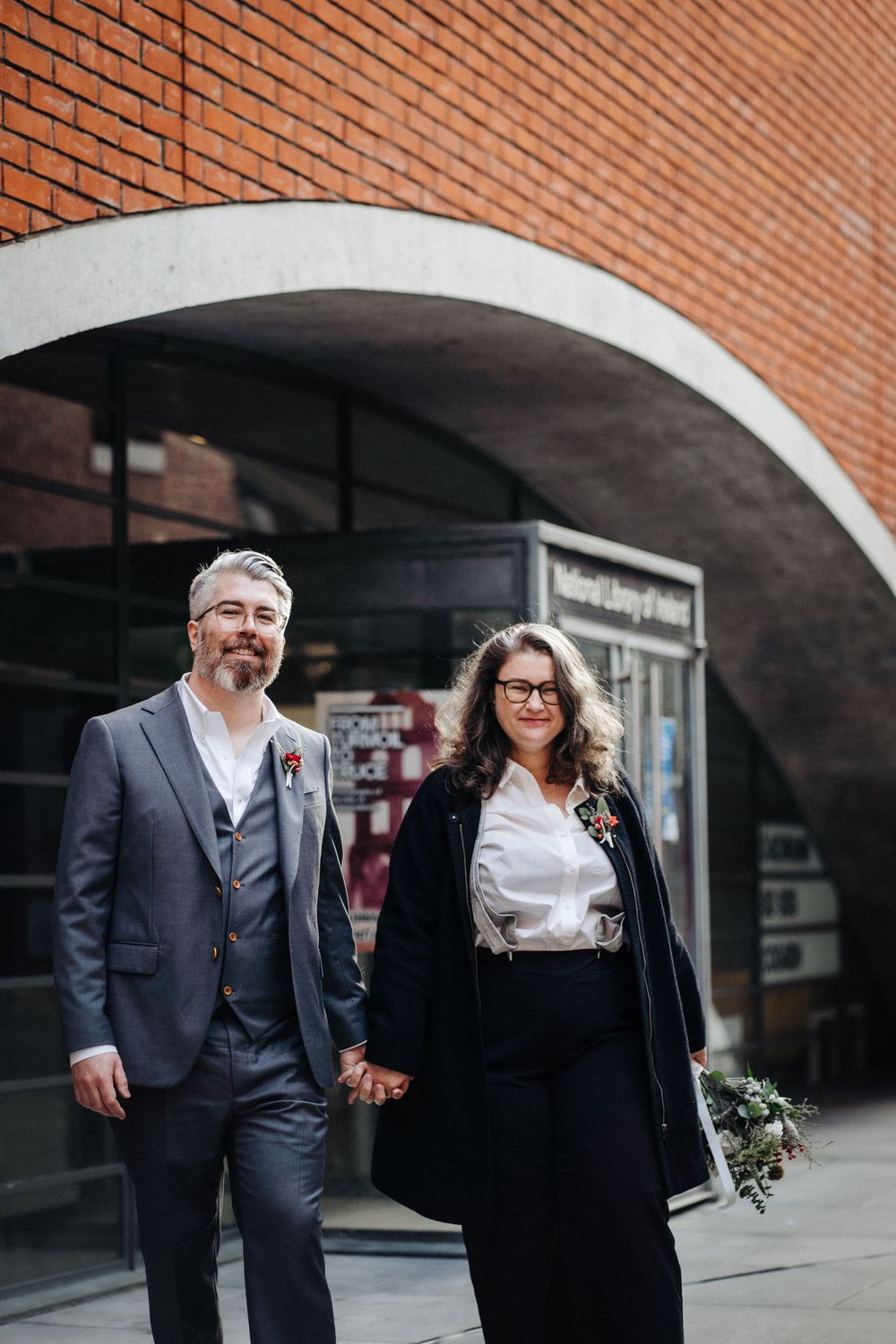 A stylish couple holds hands under a modern brick archway.