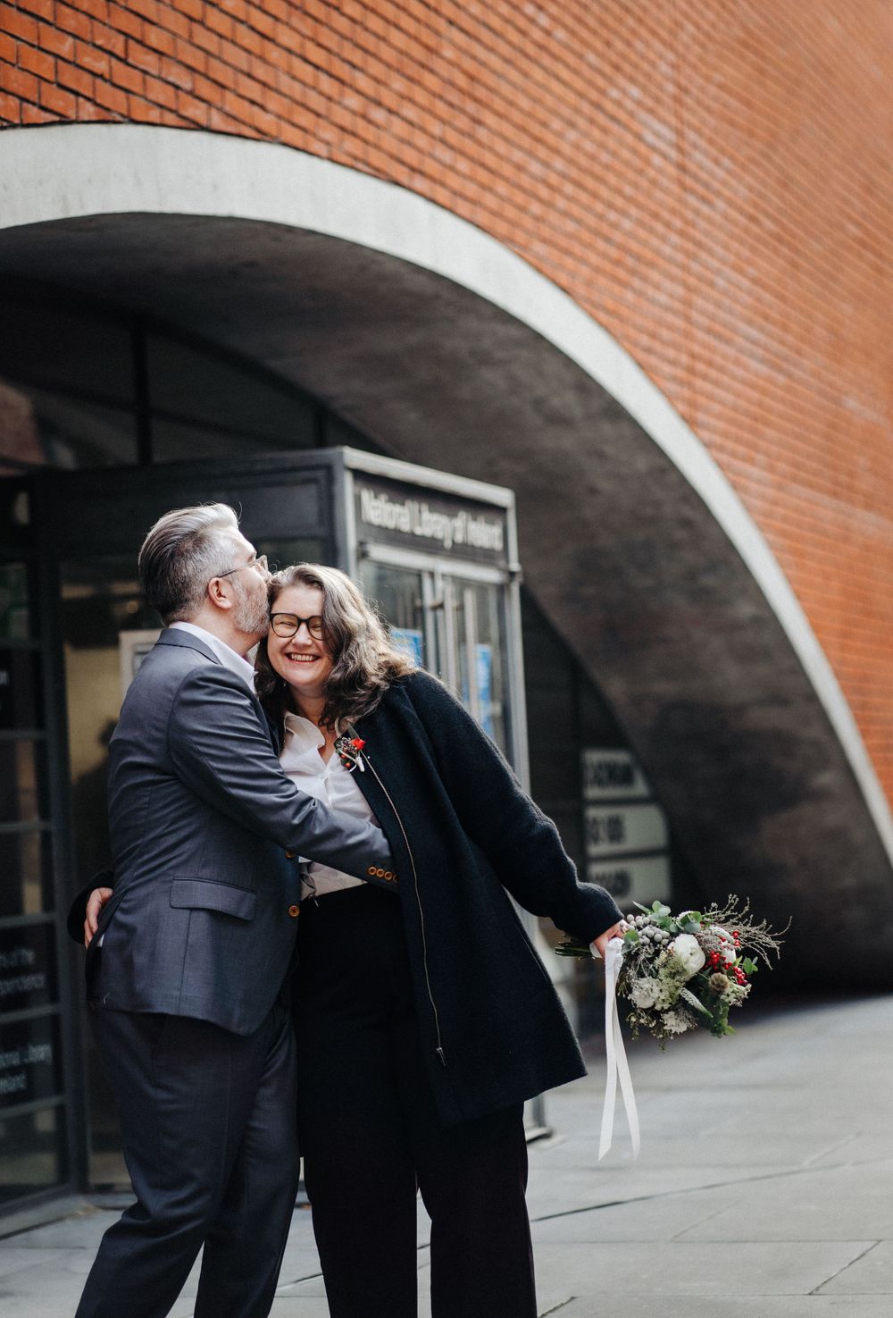 A romantic couple embraces under a brick archway while holding a floral bouquet in an urban setting.