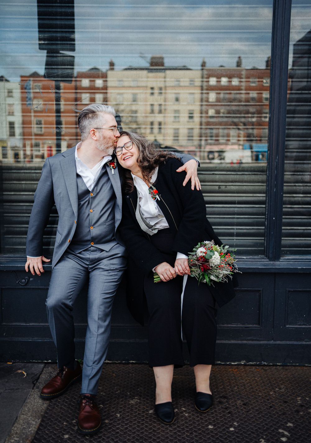 A couple shares a tender moment against a black wall with vintage buildings in the background.