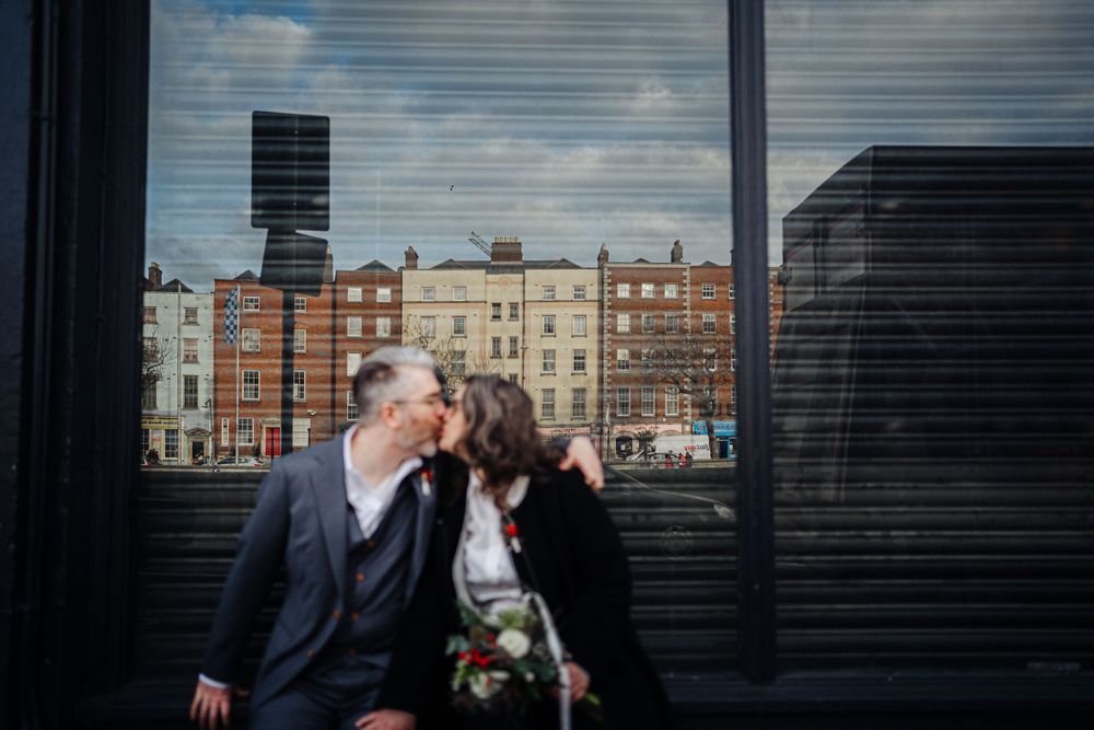 A romantic couple sits together against an urban backdrop of historic brick buildings.