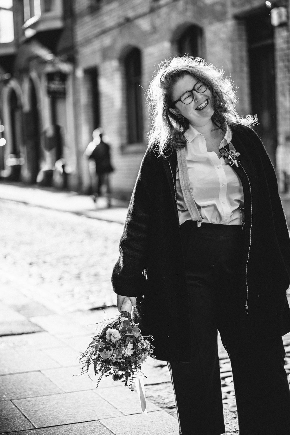 A joyful person in black attire holds a bouquet while walking down a cobblestone street.