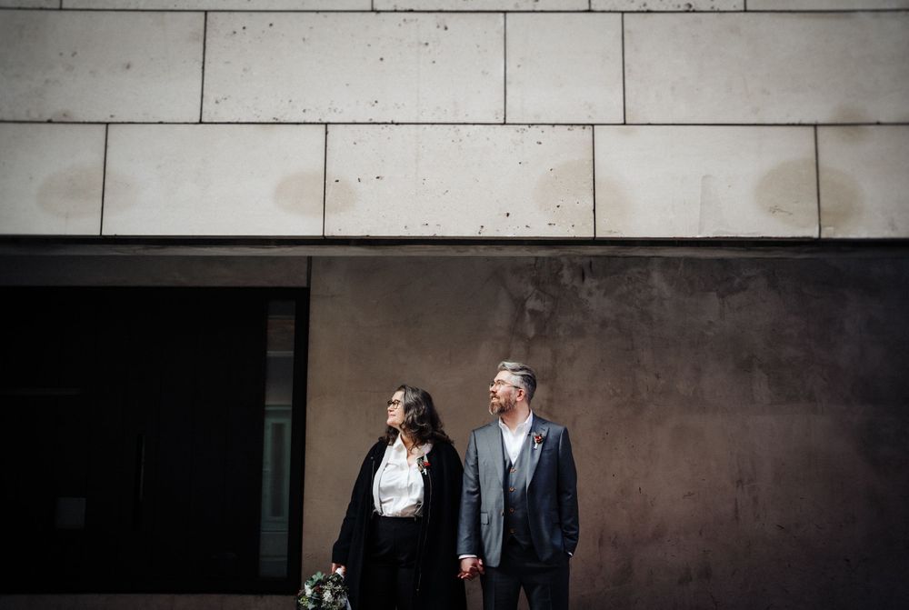 A couple stands against a modern concrete wall in an urban setting.