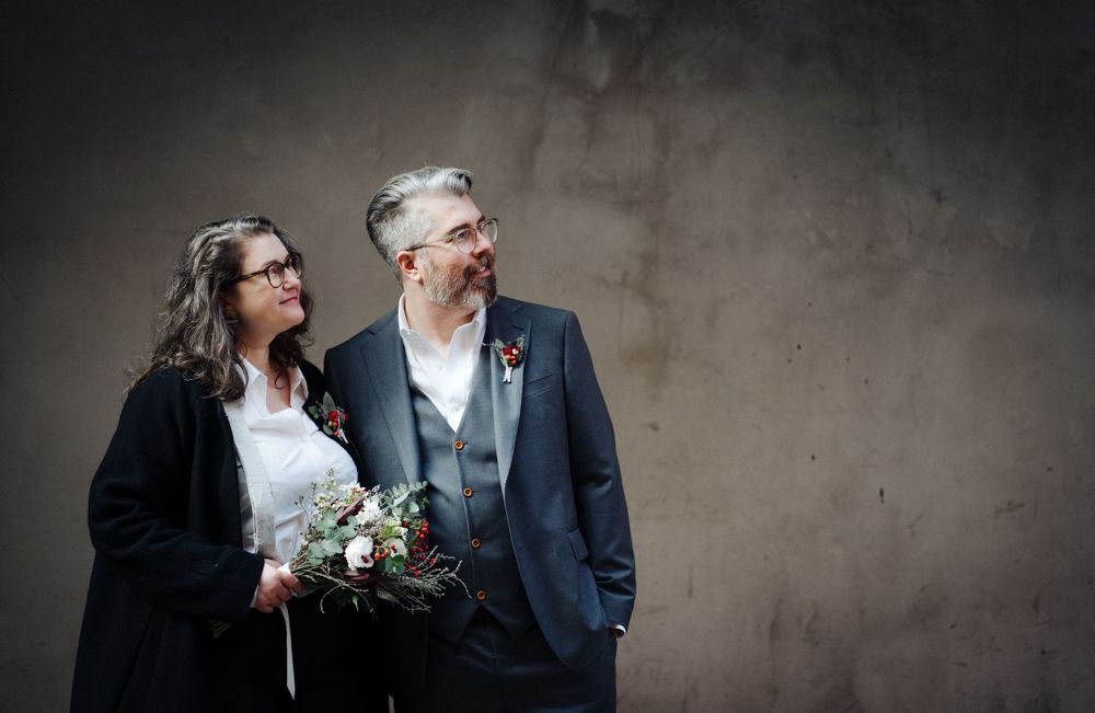 A couple poses against a textured grey wall while holding a wedding bouquet.