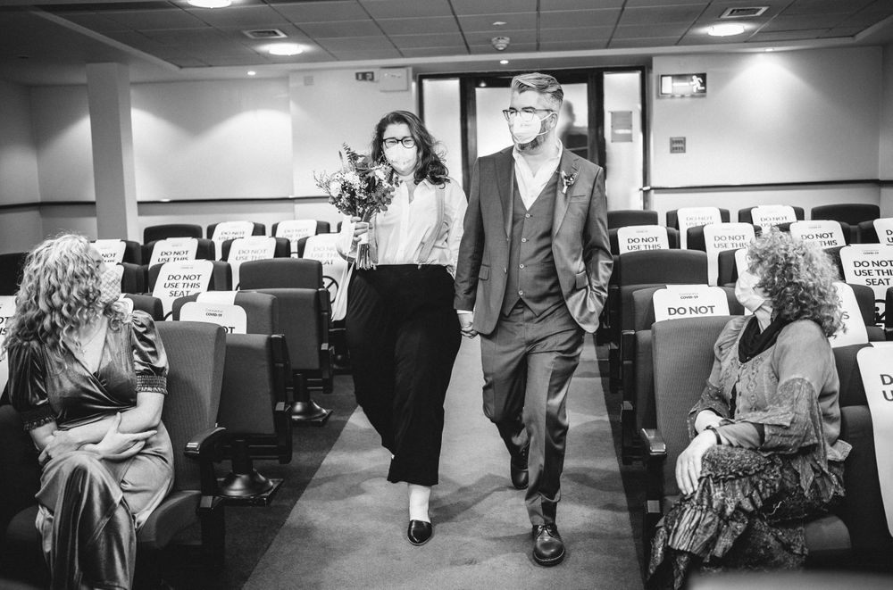 A couple walking down an aisle while guests look on in a black and white indoor ceremony setting.