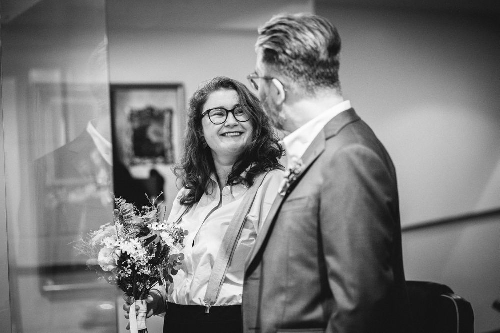 A loving couple gazes at each other while holding a floral bouquet in a hallway setting.