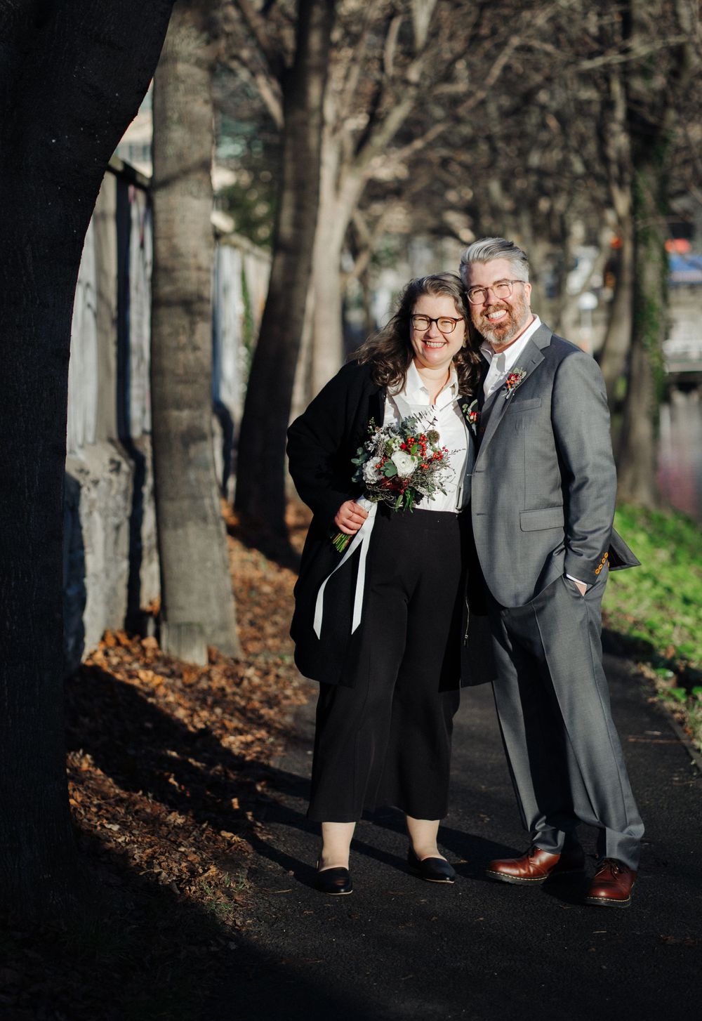 A couple poses together outdoors on a tree-lined path, dressed elegantly with a bouquet.