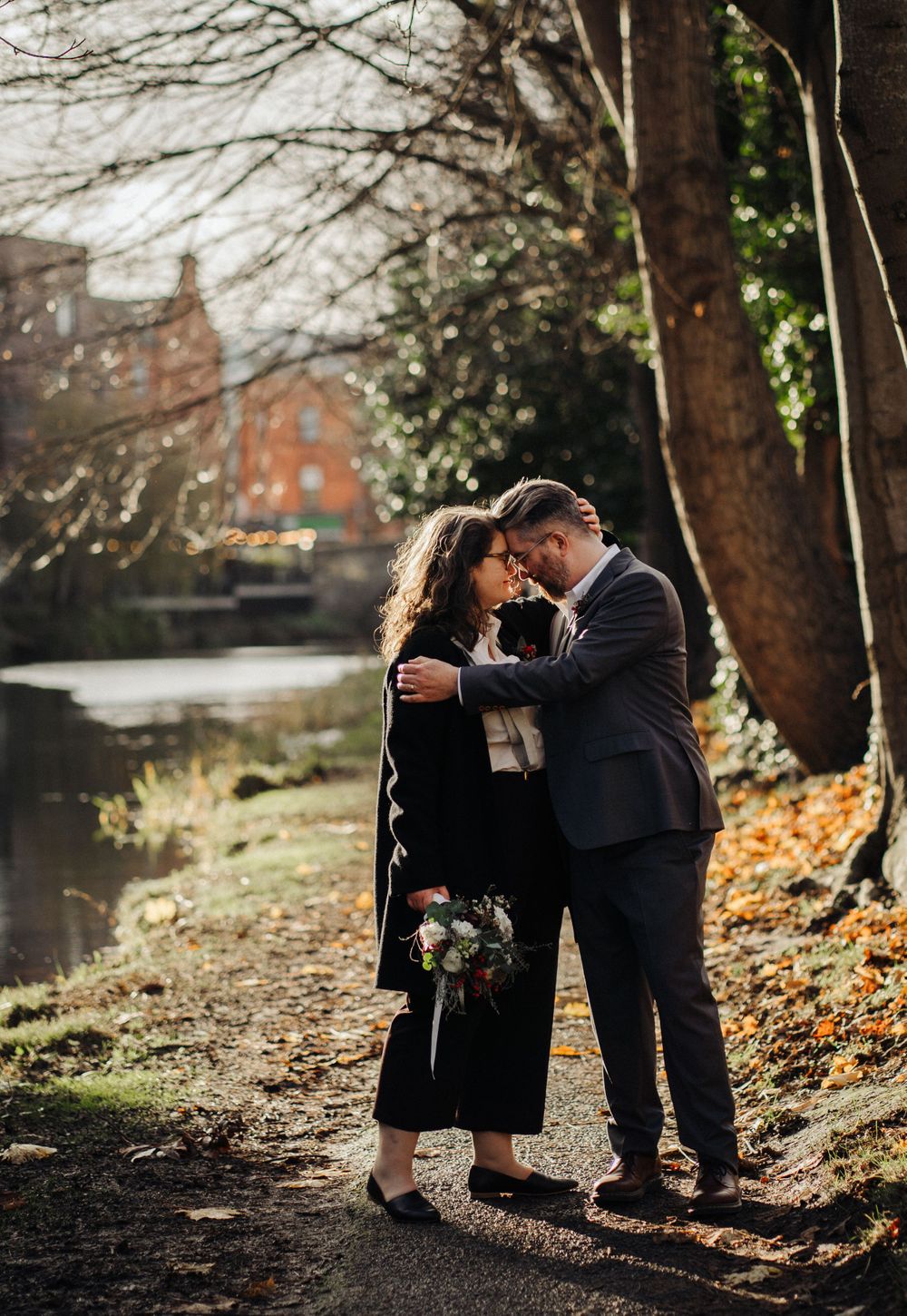 A romantic moment captured by a pond with autumn leaves and trees in the background.