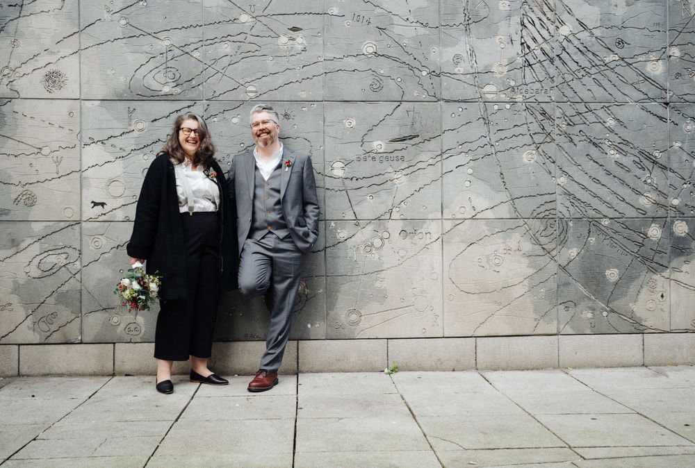 Couple poses against abstract concrete wall mural in formal attire.