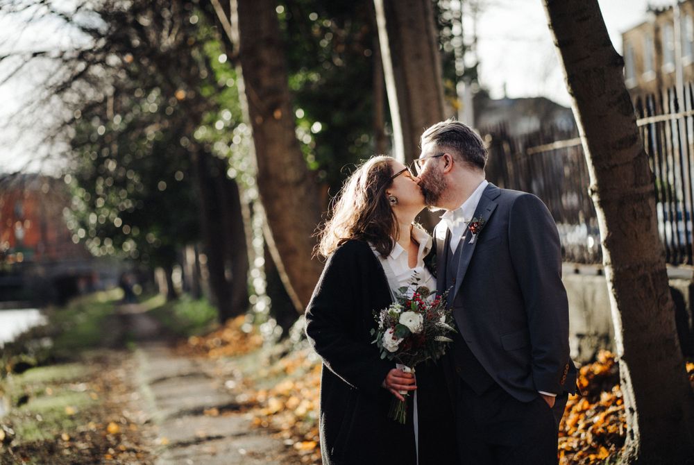 A couple shares an intimate moment on a tree-lined path with fallen autumn leaves.