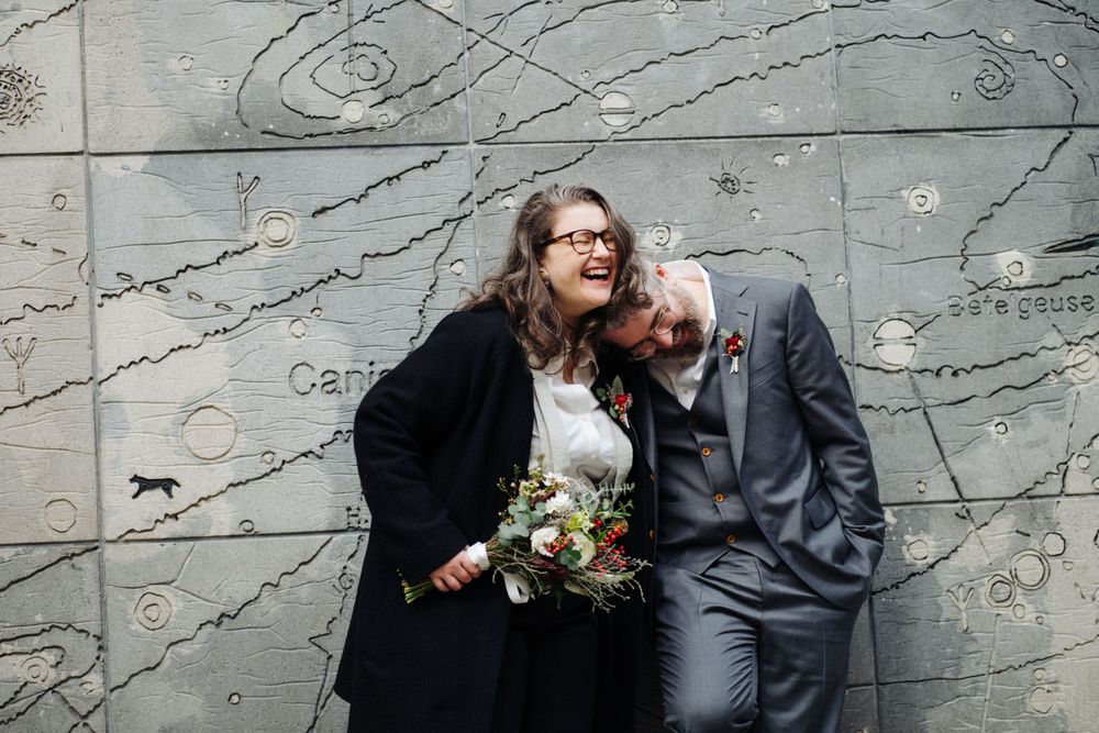 A couple poses against a textured gray wall while holding a wedding bouquet.