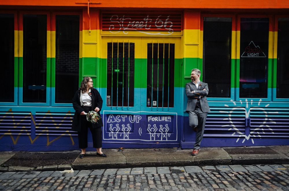 Two people pose in front of a colorful rainbow-striped building exterior.