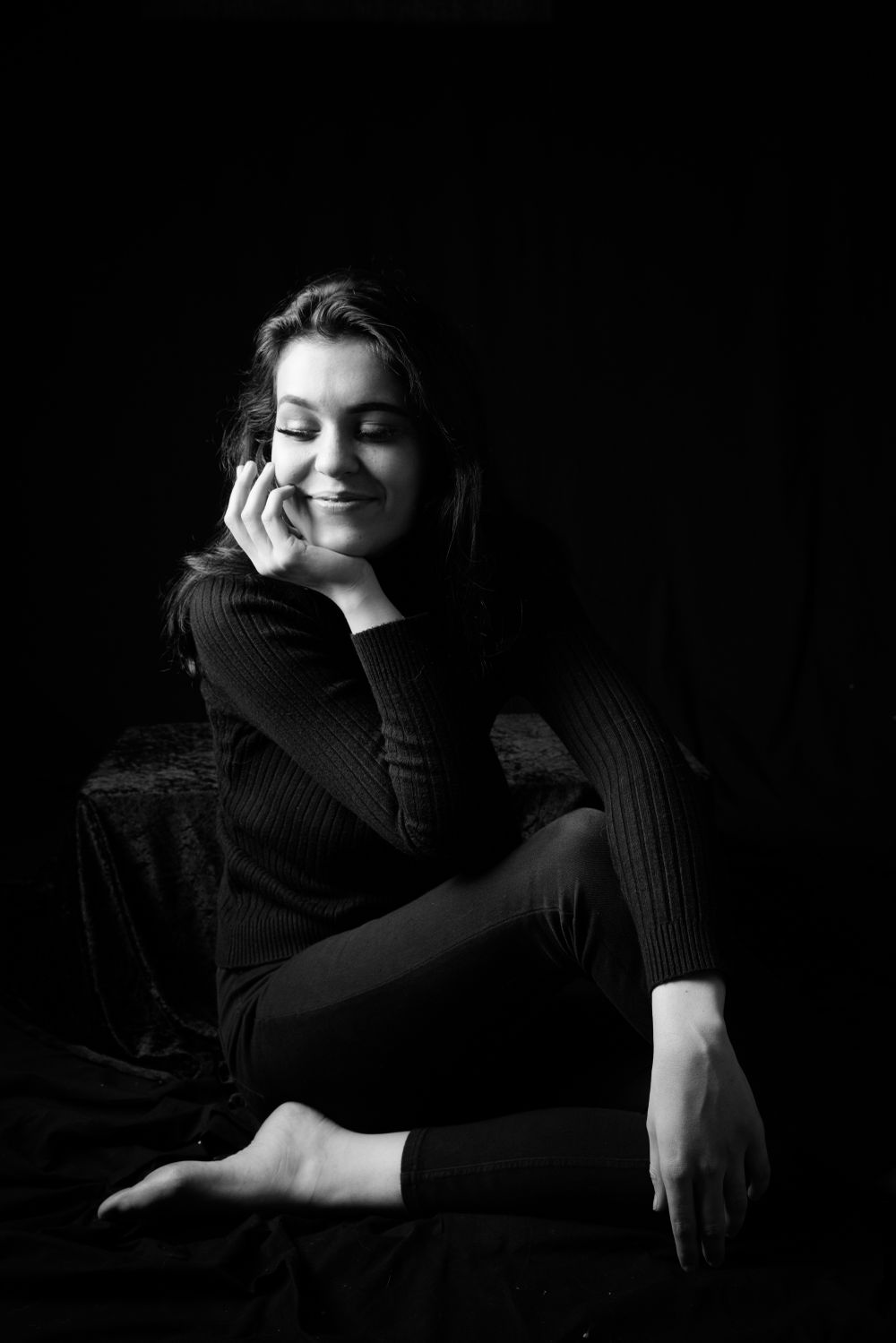 Artistic black and white portrait showing a person in dark sweater resting on furniture against black background.