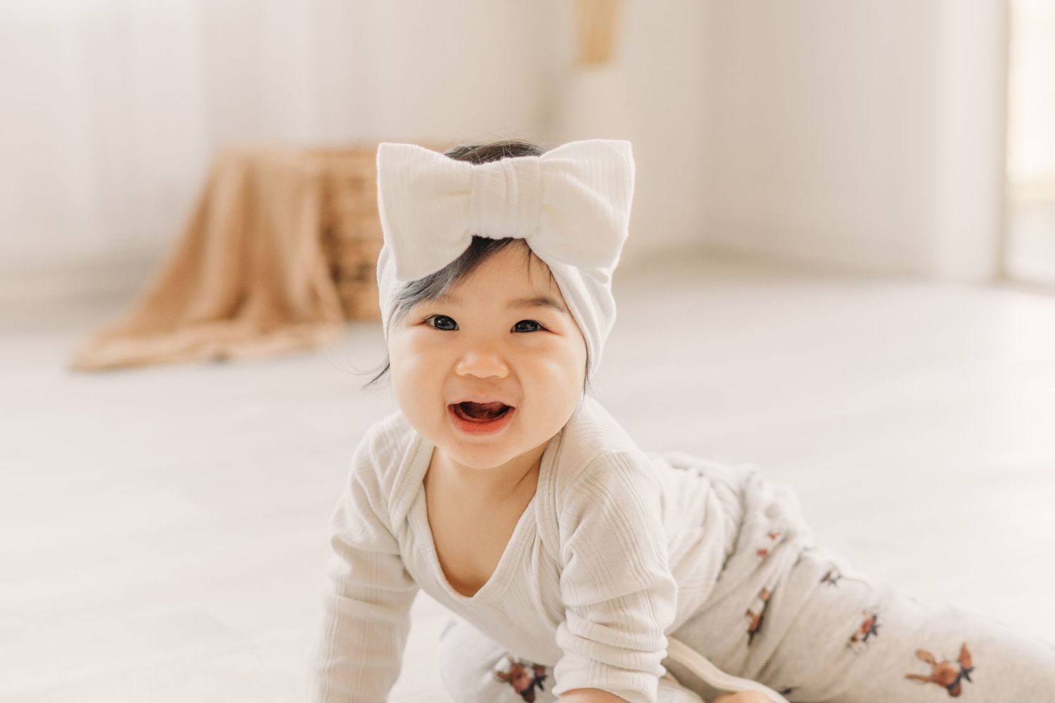 Baby in white outfit with bow crawling happily on light floor.