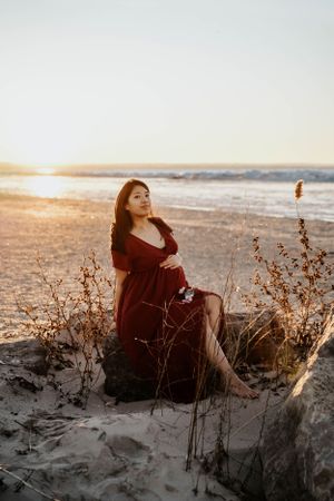 Maternity photo of woman in red dress sitting on beach dunes at golden hour.